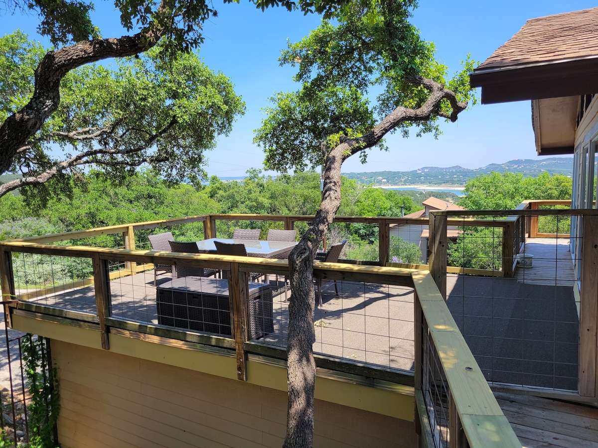A spacious deck is surrounded by lush greenery, offering views of the lake in the distance. Outdoor seating is arranged around a coffee table, and sturdy railings provide safety while enhancing the open atmosphere.
