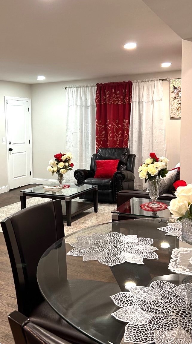 A welcoming living area features a comfortable black armchair and a glass coffee table adorned with a flower arrangement. Sheer curtains allow light to filter in, while a dining table with an intricate lace tablecloth provides a cozy gathering space. Decorative elements add a touch of elegance.