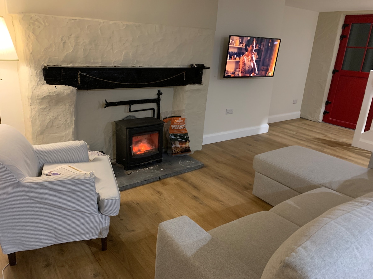 A cozy living area features a stone fireplace with a fire glowing softly. Nearby, a flat-screen television is mounted on the wall, and comfortable seating includes two light-colored armchairs and a sectional sofa. The room has warm wood flooring and neutral wall tones.