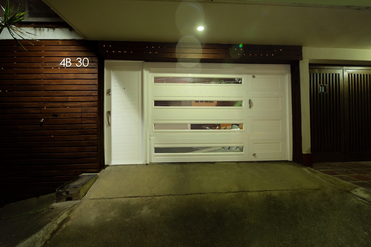 The entrance features a modern garage door with horizontal glass panels, allowing partial visibility inside. The lit area highlights the sleek design of the door, which is flanked by a wooden panel that adds texture to the exterior.