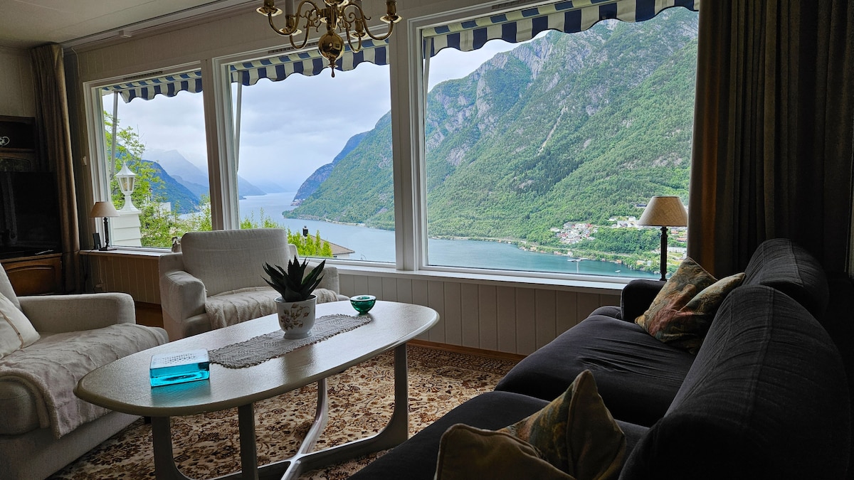 A comfortable living area features a large window with striped awnings, offering expansive views of Sørfjorden and surrounding mountains. A coffee table with a decorative plant sits between a light-colored sofa and a dark couch, creating a cozy space for relaxation.