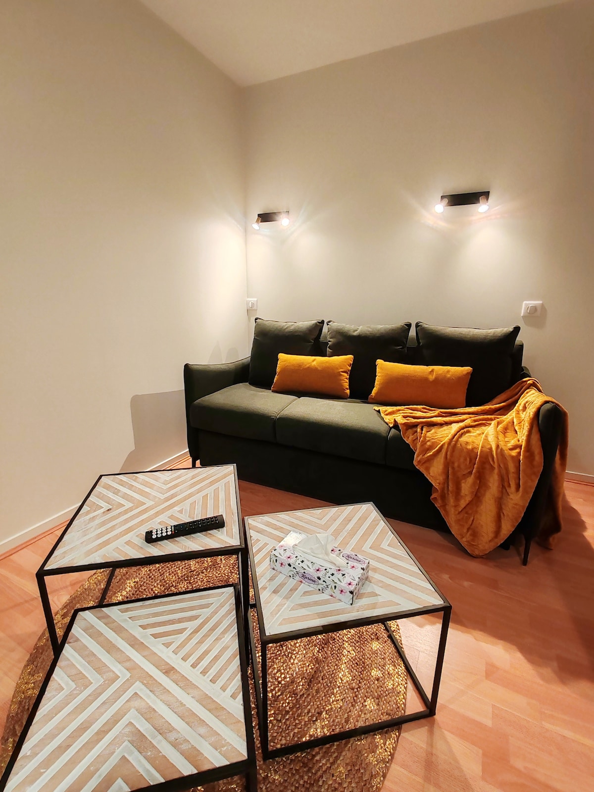 A cozy seating area is highlighted by a dark green sofa adorned with yellow cushions. A soft, golden throw is draped casually over the arm. Multiple contemporary coffee tables, featuring geometric patterns, complement the warm wood flooring.