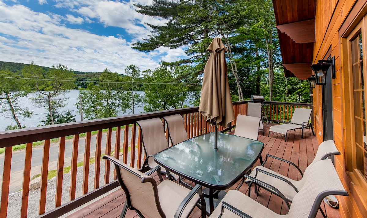A wooden balcony features a glass-topped table surrounded by six cushioned chairs. An umbrella provides shade, and the view encompasses trees and a lake under a partly cloudy sky.