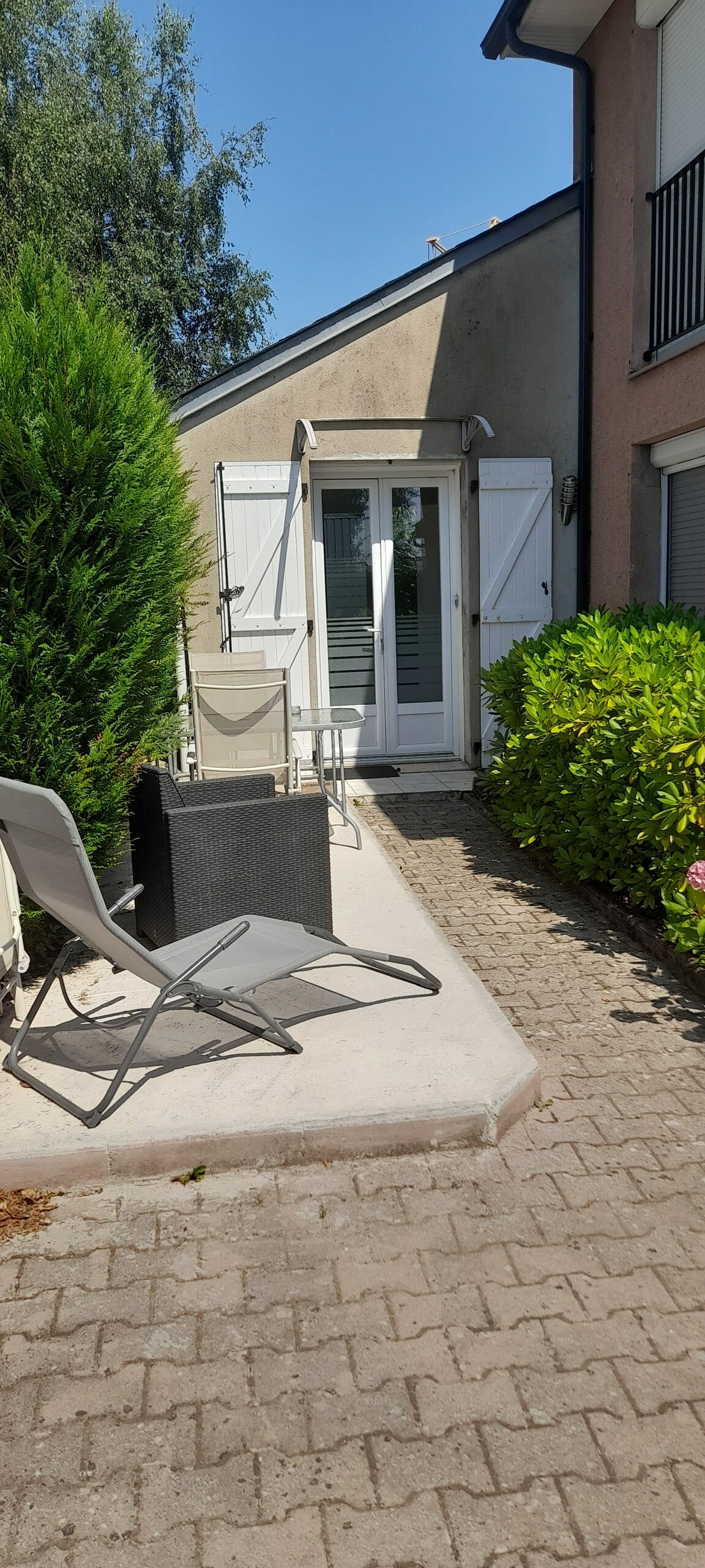 A patio area is visible beside the building, featuring two lightweight chairs and a small table. Lush greenery lines the path, including a hedge and flowering bushes. White shutters frame an open door, allowing light to enter the interior space.