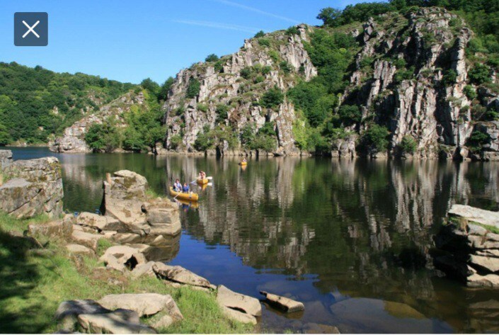 A calm river scene is depicted, with smooth rocks lining the shore. Canoes are seen gliding across the water, reflecting the surrounding greenery and rocky cliffs. The landscape is enhanced by clear blue skies and lush foliage, creating a serene outdoor environment.