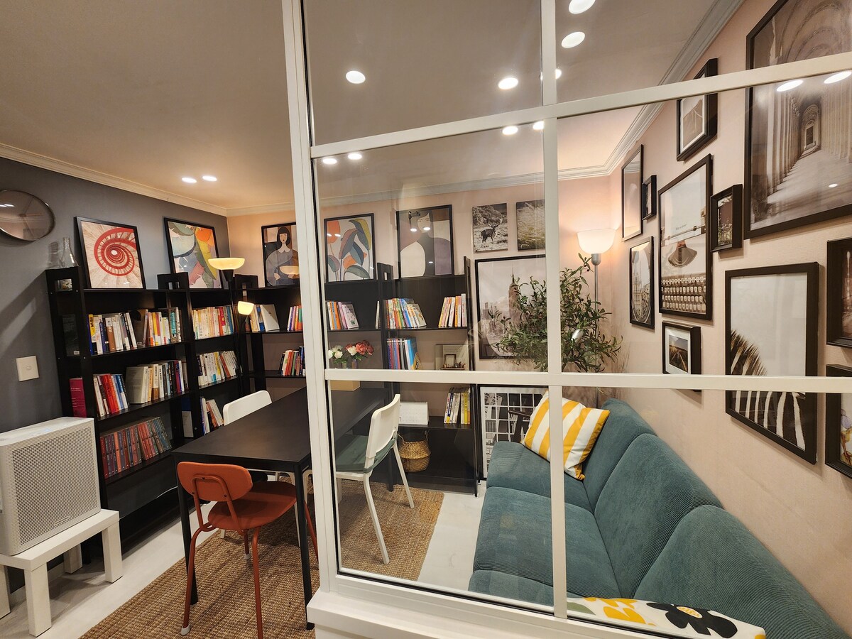 A comfortable living area is shown, featuring a spacious green sofa and an inviting coffee table. A bookshelf filled with books lines one wall, while various framed artworks decorate the surrounding space. Soft lighting enhances the cozy atmosphere.