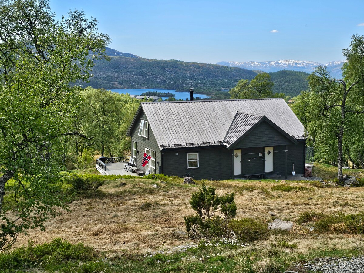 A modern cabin is set against a scenic mountain backdrop, showcasing a sloped roof and large windows. Surrounding greenery includes trees and shrubs, with a driveway leading to the entrance. A glimpse of water can be seen in the distance, framed by the natural landscape.