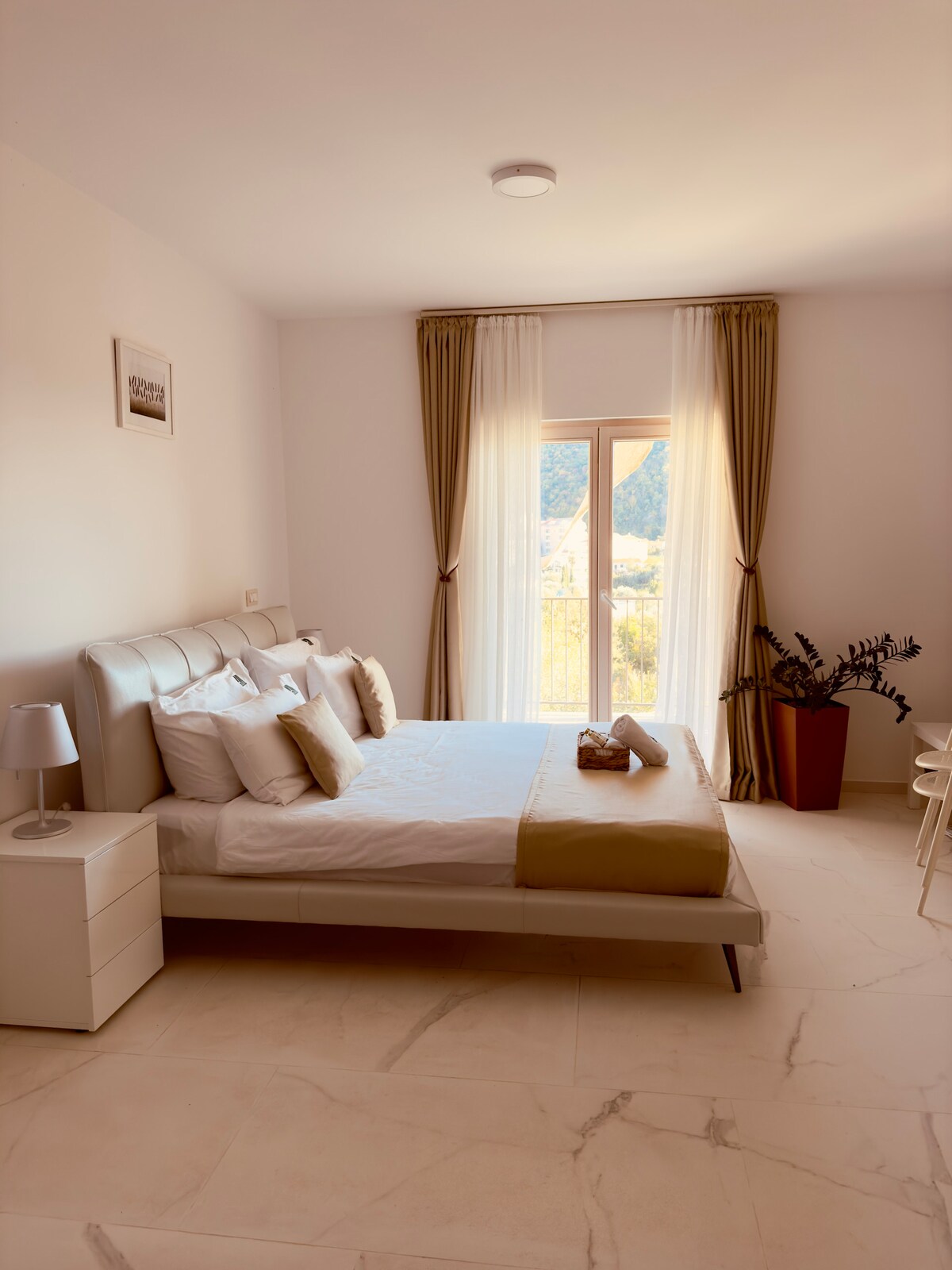 A bright and spacious sleeping area features a comfortable double bed with neutral-toned bedding. A softly lit bedside lamp rests on a nightstand. Floor-to-ceiling windows allow natural light to fill the room, complemented by flowing curtains and a decorative plant in a stylish pot.