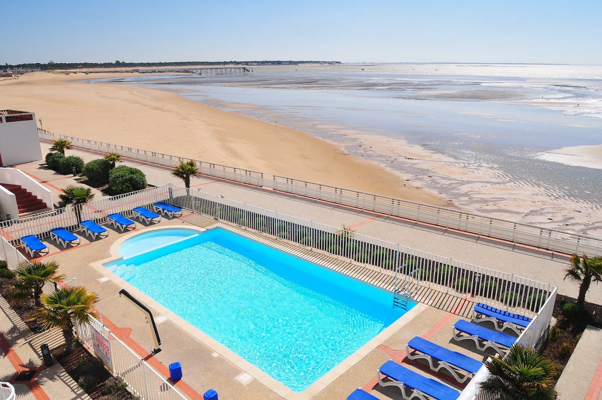 A clear view of a sparkling blue swimming pool is framed by sun loungers. The beach stretches beyond with soft, golden sand and water meeting the horizon. Lush greenery and paving surround the pool area, providing a serene setting directly adjacent to the seaside.