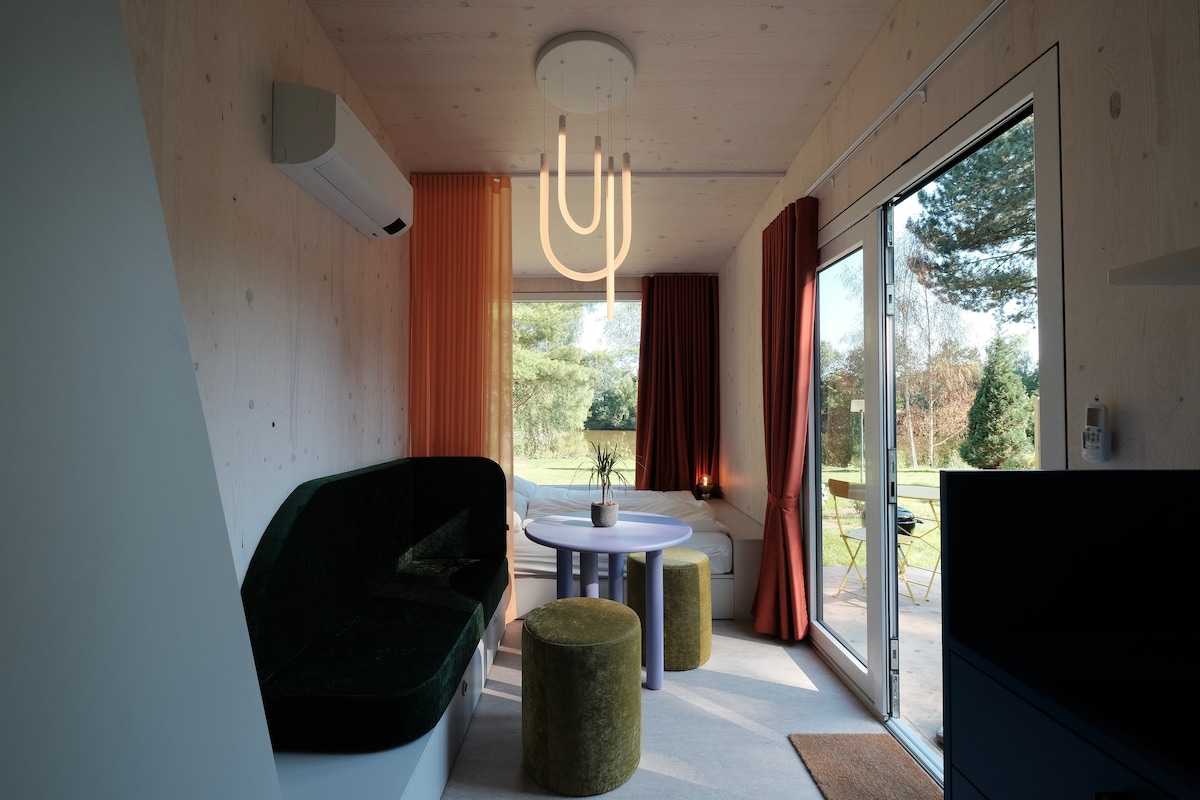 A modern living space features a uniquely shaped green sofa and a round dining table surrounded by two plush stools. Large glass doors allow views of the outdoor greenery, while soft orange and deep red curtains frame the windows, complementing the lighting fixture above.