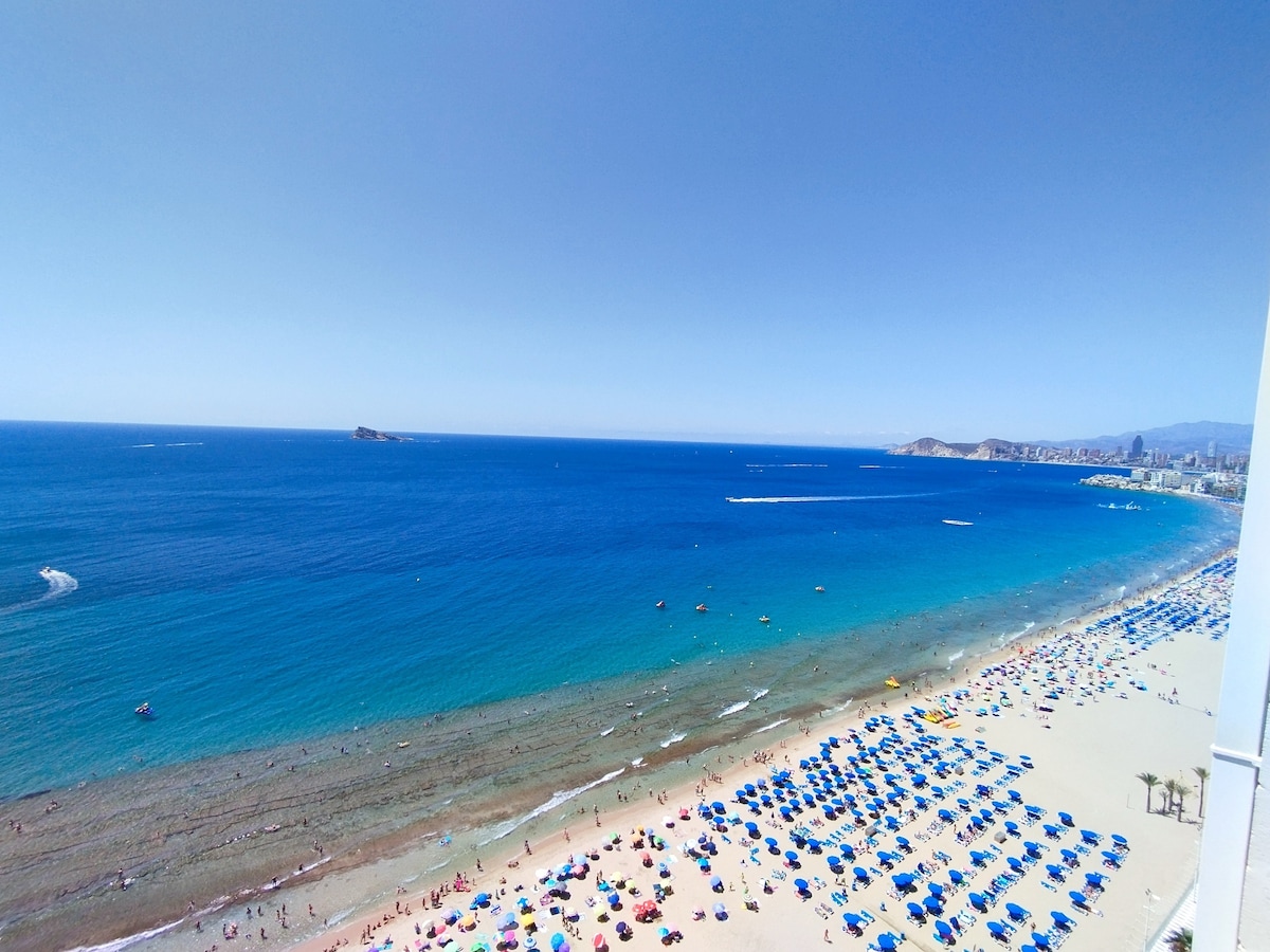 A panoramic view captures the vibrant beach of Levante, filled with colorful umbrellas and sunbathers. The clear blue waters gently lap against the golden sand, while distant boats traverse the horizon under a bright blue sky, enhancing the coastal ambiance of Benidorm.