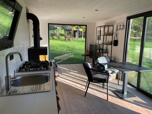 An interior space features large glass doors that offer views of the green outdoor landscape. A modern kitchenette includes a stainless steel sink and a gas stove. Seating consists of a black wicker chair and a sleek table, while sunlight softly illuminates the wooden floor.
