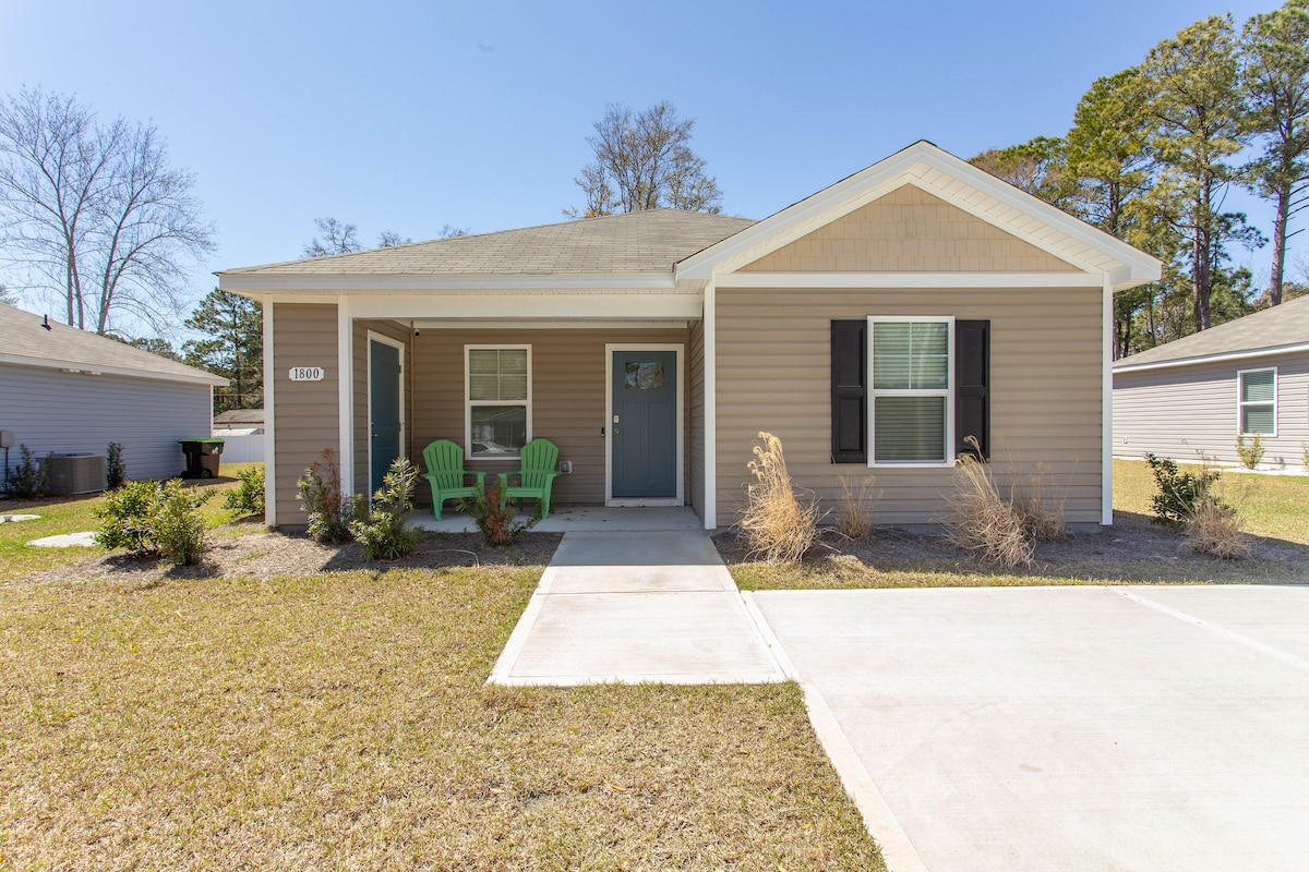 The exterior of a single-story home features a light brown facade with black shutters and a welcoming front porch. Two green chairs are positioned on the porch, surrounded by a well-maintained lawn and landscaping. A concrete driveway leads to the entrance.