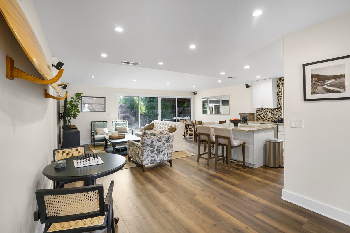 An open-concept living space is illuminated by recessed lighting, featuring a mix of seating arrangements including a floral-patterned sofa and a dining table. A chess set is positioned on a small table, complementing the modern kitchen visible in the background.
