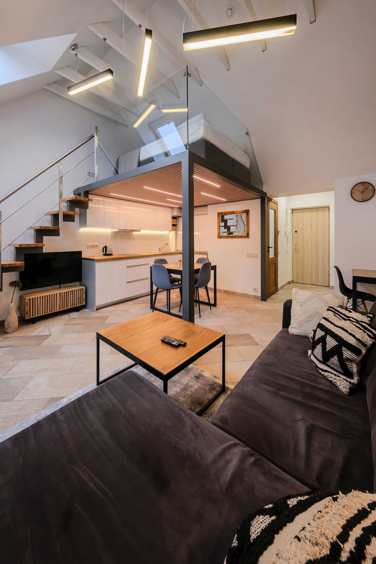 An open-concept studio features a modern design with a comfortable dark sofa facing a wooden coffee table. A spiral staircase leads to an upper entresol area. The kitchen, equipped with contemporary appliances, is visible in the background, with lighting that highlights the space.