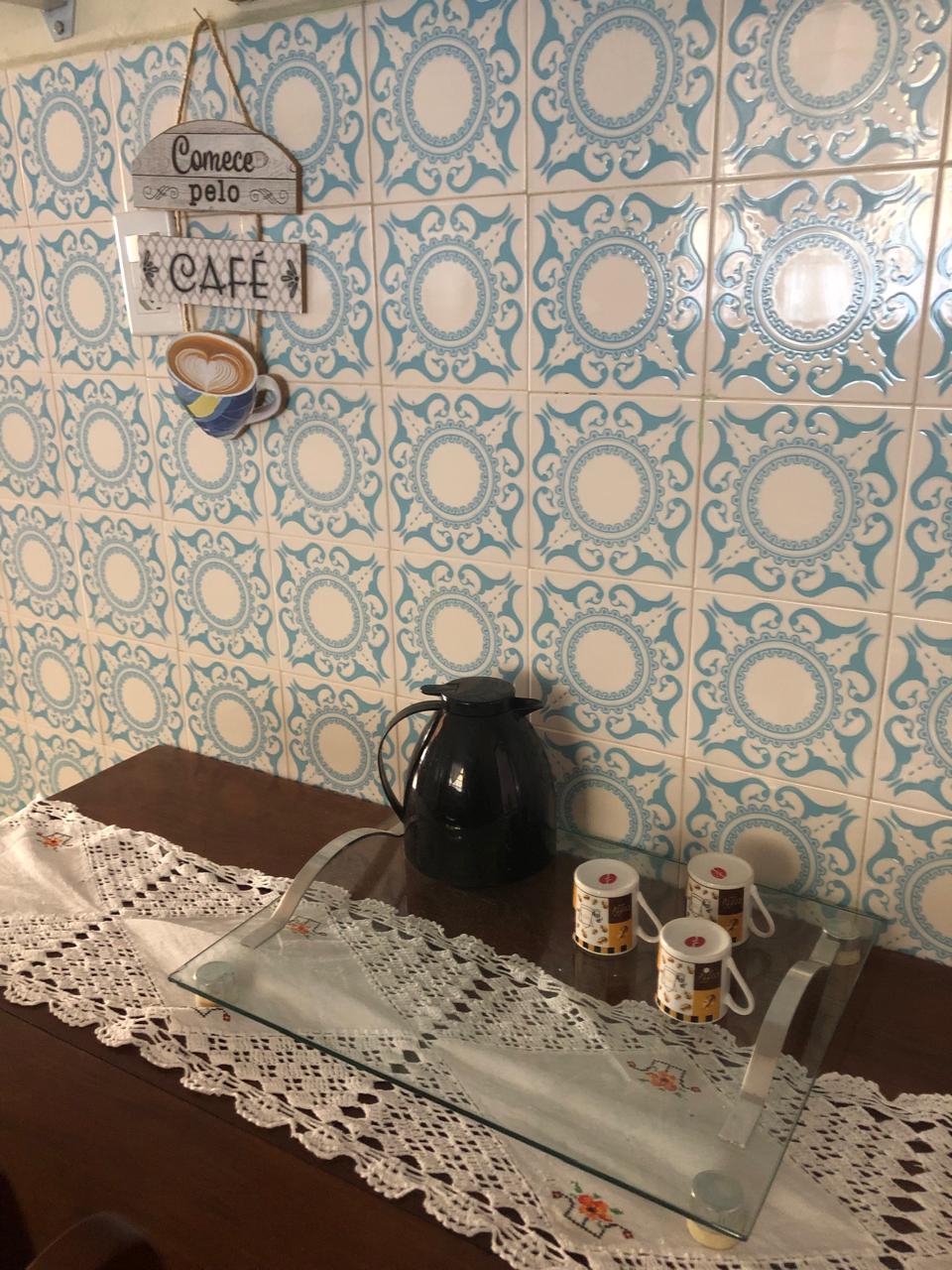 A wooden table is adorned with a delicate lace runner, featuring a glass tray at the center. Several coffee mugs, each showcasing unique patterns, are positioned beside a sleek black coffee pot. A decorative sign hanging above adds charm to the backdrop of blue patterned tiles.