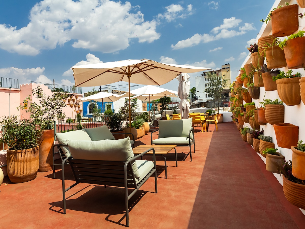 The rooftop space features comfortable seating arranged under large umbrellas, with greenery provided by potted plants lining the walls. A bright blue sky and scattered clouds hover above, creating a serene environment for relaxation.
