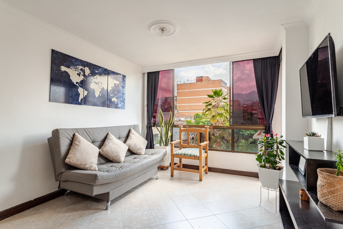 A bright living area is showcased with a comfortable gray sofa and an accent chair featuring natural wood. Green plants are arranged near the large window, which offers city views. The wall is adorned with a world map, enhancing the space's visual interest.