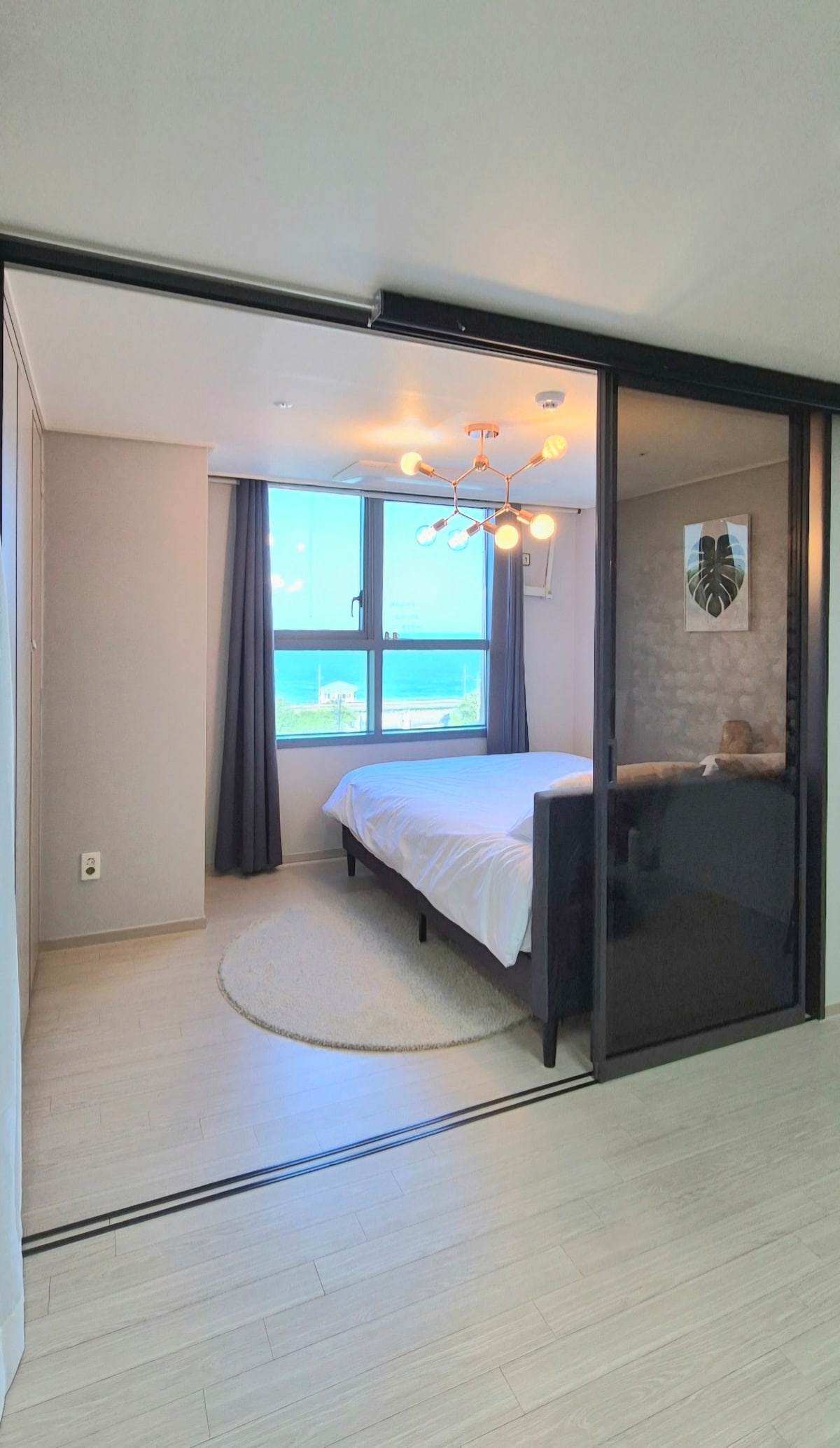 A serene sleeping area is framed by a sliding glass door, featuring a spacious bed and a stylish light fixture above. Large windows offer views of the ocean, with light filtering through dark curtains, and a round rug adds a touch of comfort to the floor.