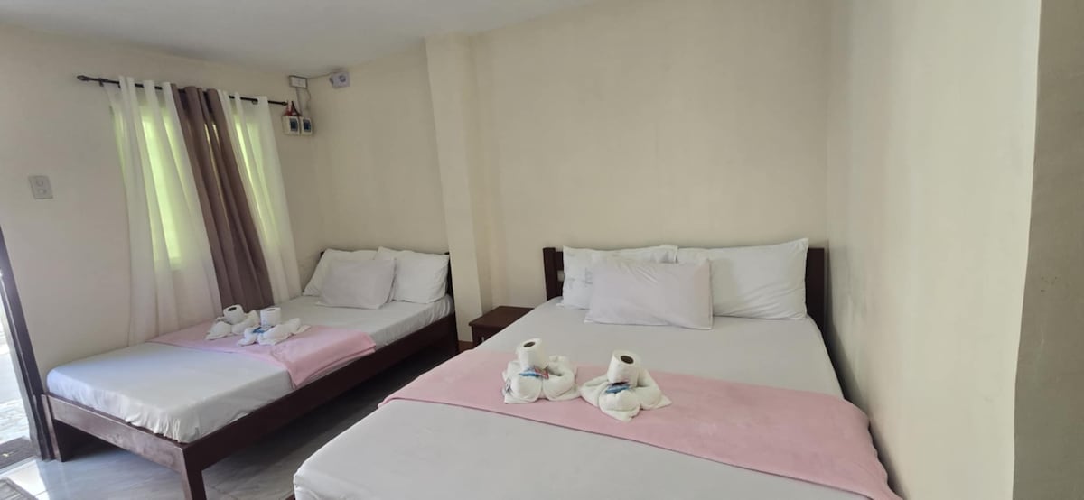 A welcoming room is furnished with two spacious beds adorned with light pink blankets. Fresh towels are neatly arranged on each bed. Natural light filters through the adjacent window, partially dressed with light curtains, illuminating the neutral-colored walls.