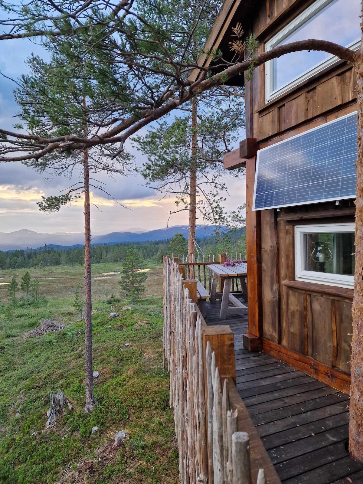 A wooden terrace extends from the cabin, surrounded by natural scenery with distant mountains visible. Solar panels are mounted on the external wall, and a table is set for outdoor dining. Tall trees frame the view, enhancing the serene atmosphere.