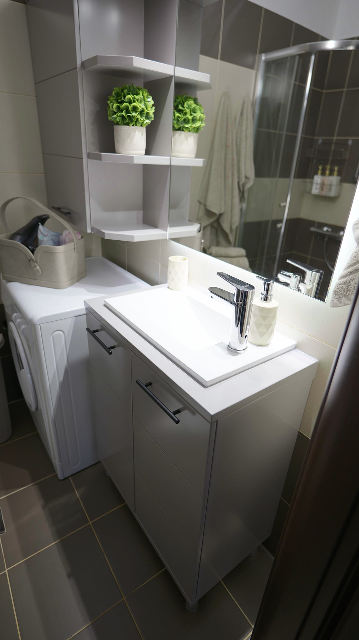 A modern bathroom is presented with a sleek vanity featuring drawers and a minimalist sink. Above the sink, a shelf holds decorative plants. A washing machine is neatly integrated into the space, with a stylish grey container placed nearby. A glass shower enclosure is visible in the background.
