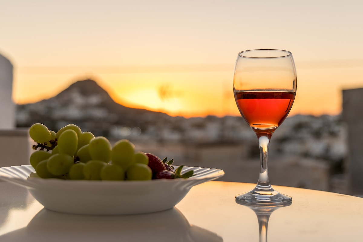 A glass of red wine is positioned alongside a plate of green grapes and strawberries. The setting sun casts a warm glow in the background, illuminating the surrounding white structures and distant hills.