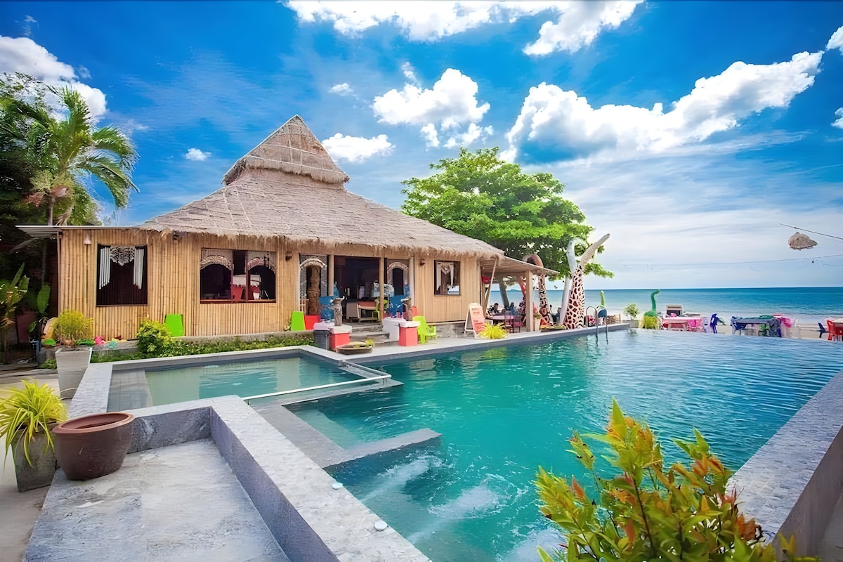 A charming beach house features a thatched roof, surrounded by a vibrant landscape. A large pool sits in the foreground, reflecting the sky, while the sandy beach and ocean are visible in the background. Colorful seating areas add to the inviting atmosphere.