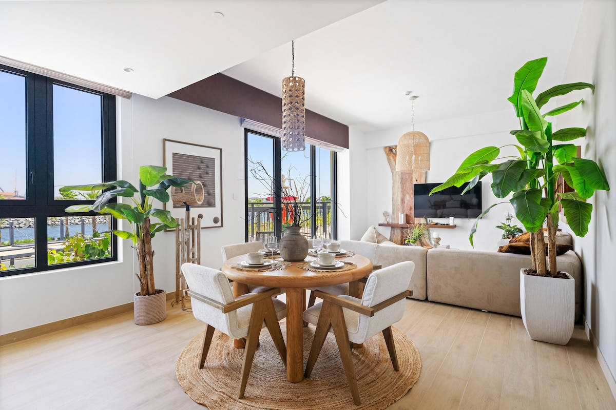 A dining area features a round wooden table with four chairs, set with plates and silverware. Tall indoor plants add a touch of greenery beside large windows that allow natural light to fill the space. The living area with a cozy sofa is visible in the background.
