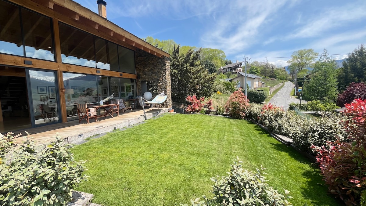 A well-maintained garden is visible in the foreground, featuring lush green grass and various shrubs. The expansive glass doors of the house open onto a spacious deck, allowing natural light to enter. Scenic views of the surrounding area and mountains are evident in the background.
