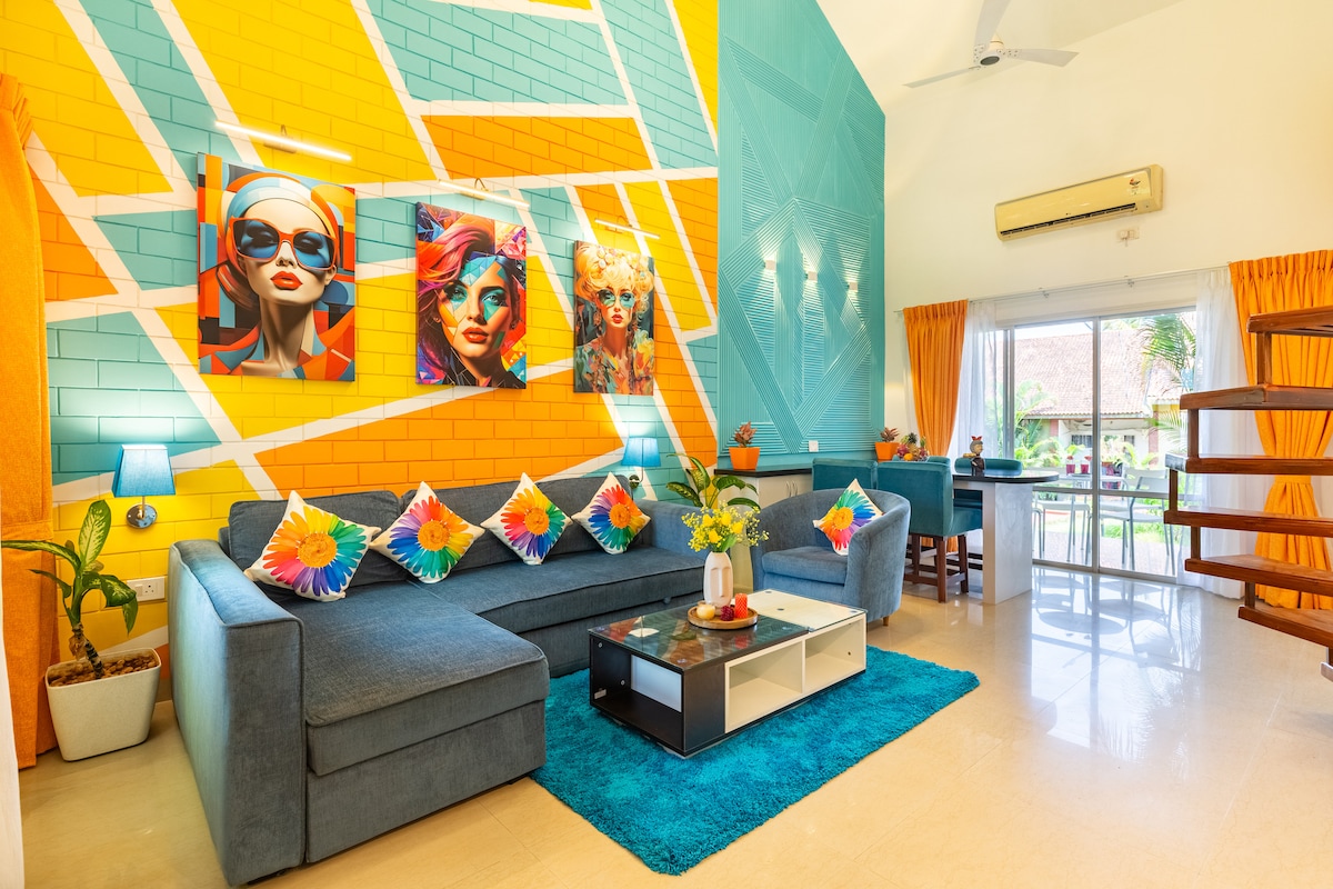 A vibrant living room features a spacious sectional sofa adorned with bright, decorative pillows. Colorful pop art adorns the walls, contrasted by bright turquoise accents. Natural light streams through large windows, illuminating the modern decor and a small dining area visible through the open layout.