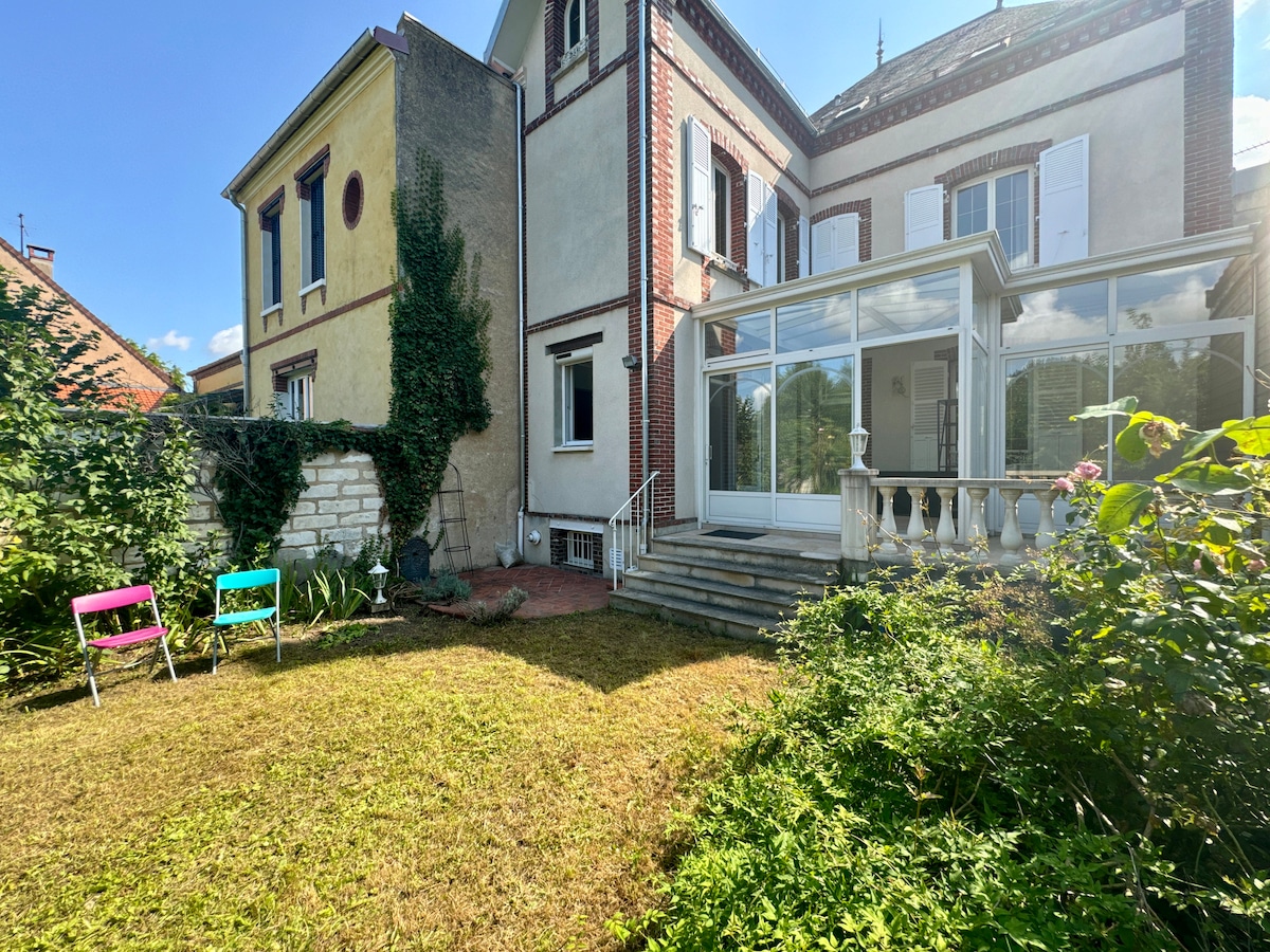 The exterior view showcases a historic residence combined with modern elements. A spacious garden area is visible, featuring green grass and colorful chairs. A bright, glass-enclosed veranda offers a welcoming space, complemented by charming brick and stone architectural details.