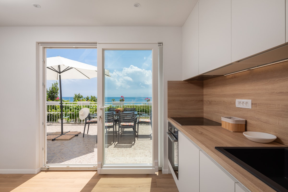 A modern, fully equipped kitchen is depicted, featuring wooden cabinetry and a sleek countertop. A glass door leads to a spacious balcony with outdoor seating and an umbrella, showcasing a serene sea view in the background.
