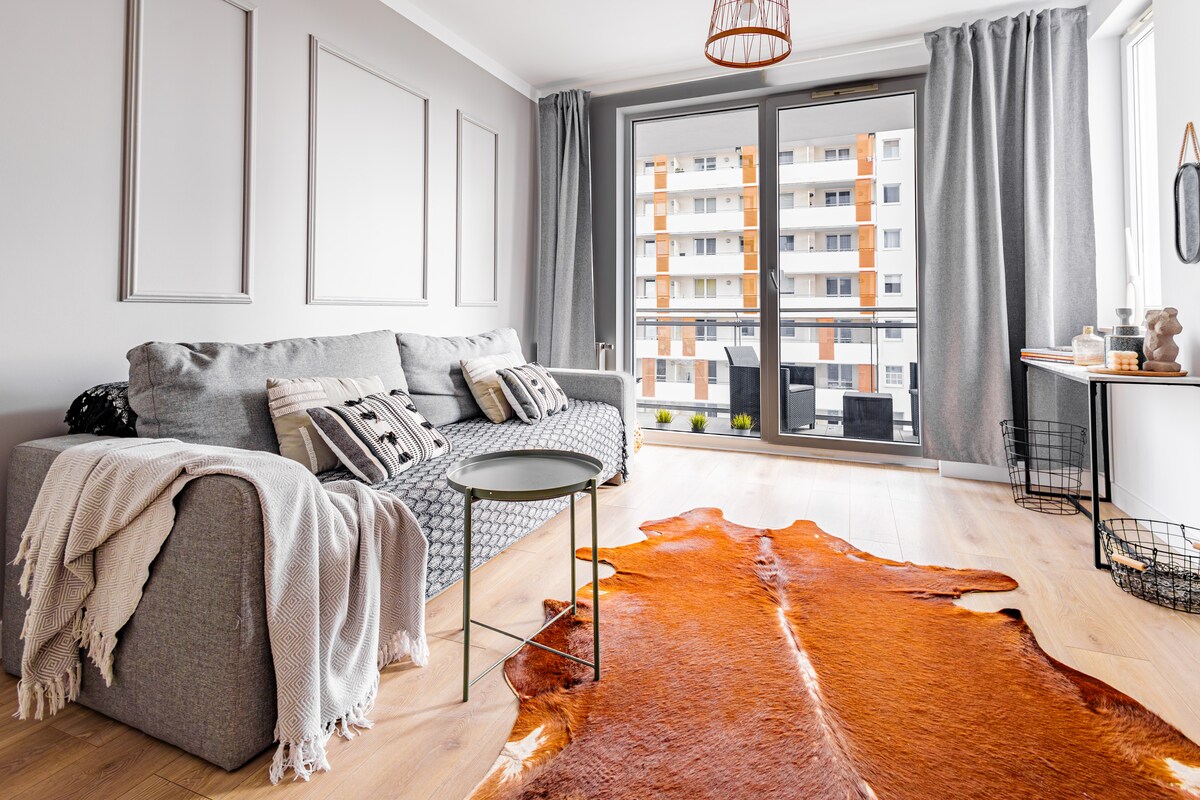 A comfortable living area showcases a light grey sofa adorned with decorative cushions. A round table is positioned beside it, and a warm-toned area rug is spread across the natural wood floor. Floor-to-ceiling glass doors provide a view of the balcony and outdoor surroundings.