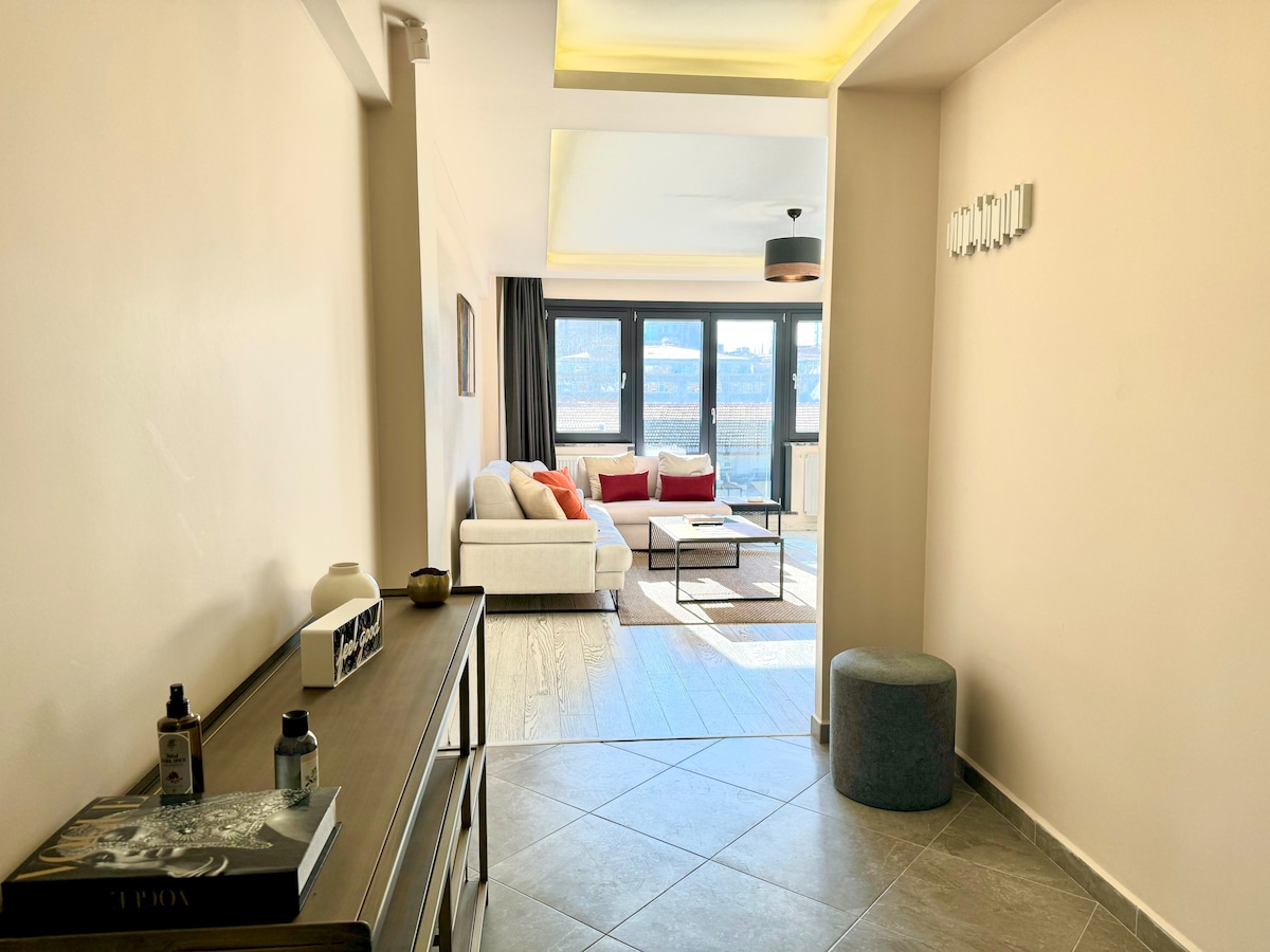 A bright entrance leads to an open living area, featuring large windows that illuminate the space. A comfortable white sofa and accent chairs are arranged around a center table, with a soft area rug beneath. Decorative elements add a welcoming touch to the room.
