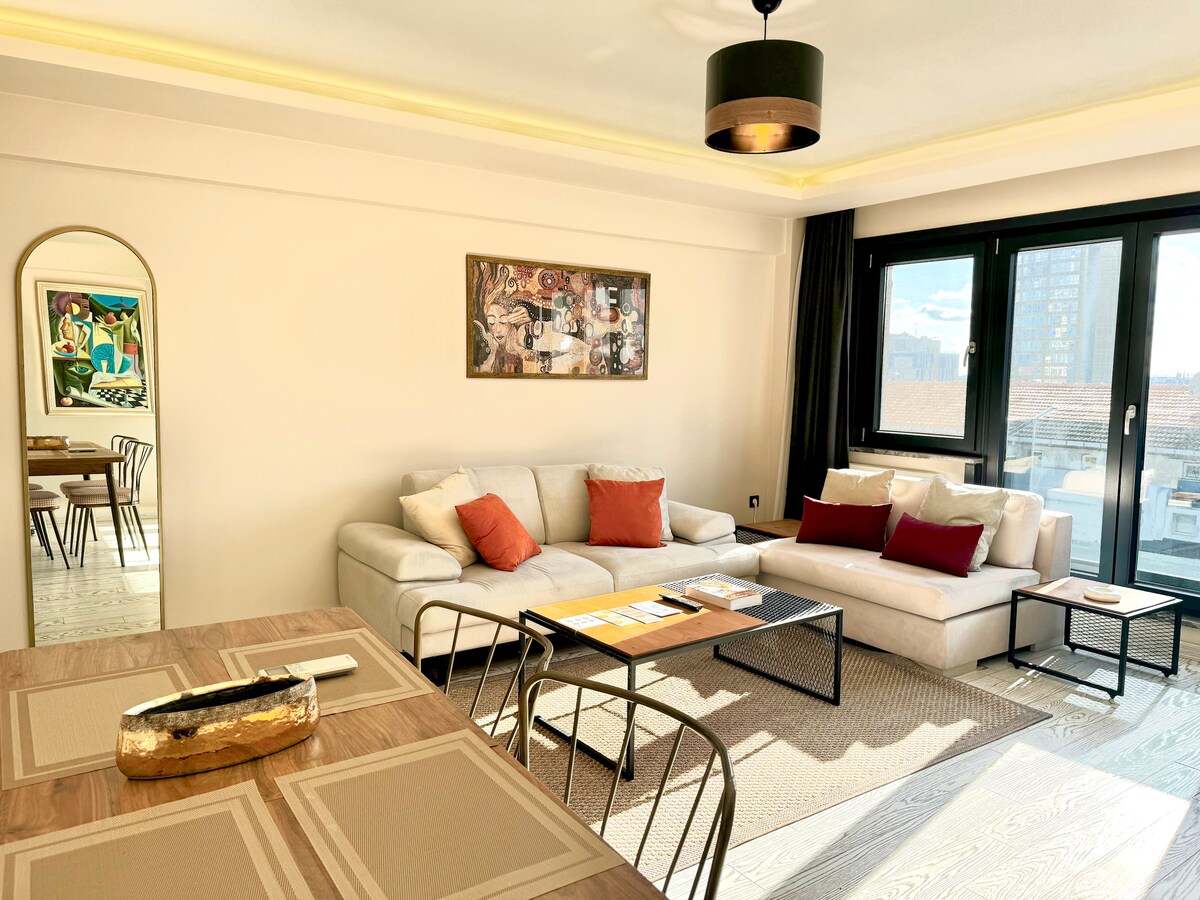 A modern living room is showcased, featuring a comfortable sofa adorned with decorative cushions and a stylish center table. A dining area with a wooden table for four is also visible. Large windows allow natural light to brighten the space, complemented by warm lighting.