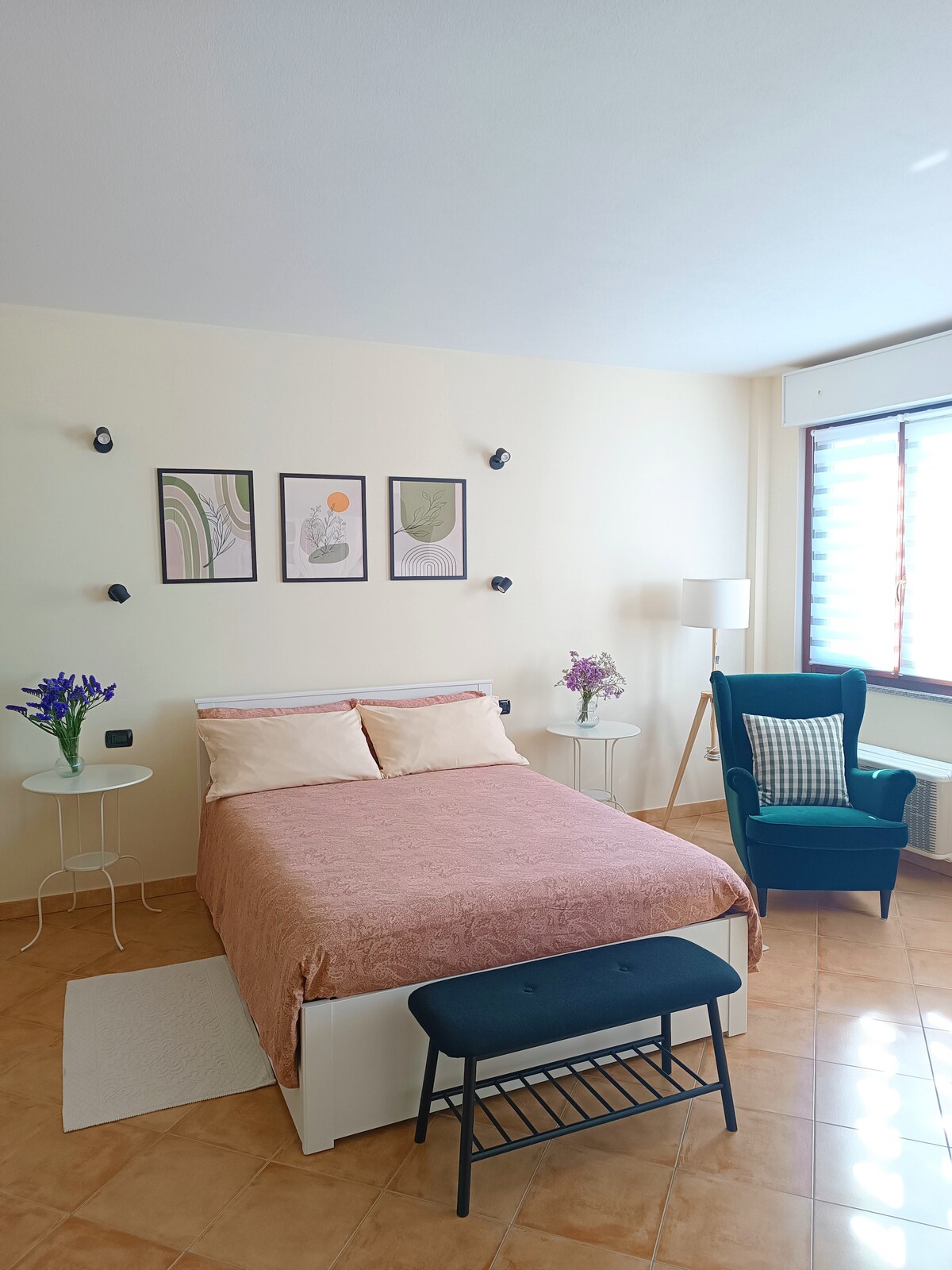 A cozy sleeping area features a spacious bed with a soft pink coverlet. Two nightstands with flowers are positioned beside the bed. A green accent chair is visible, complemented by artwork on the wall and natural light streaming through a window covered with blinds.