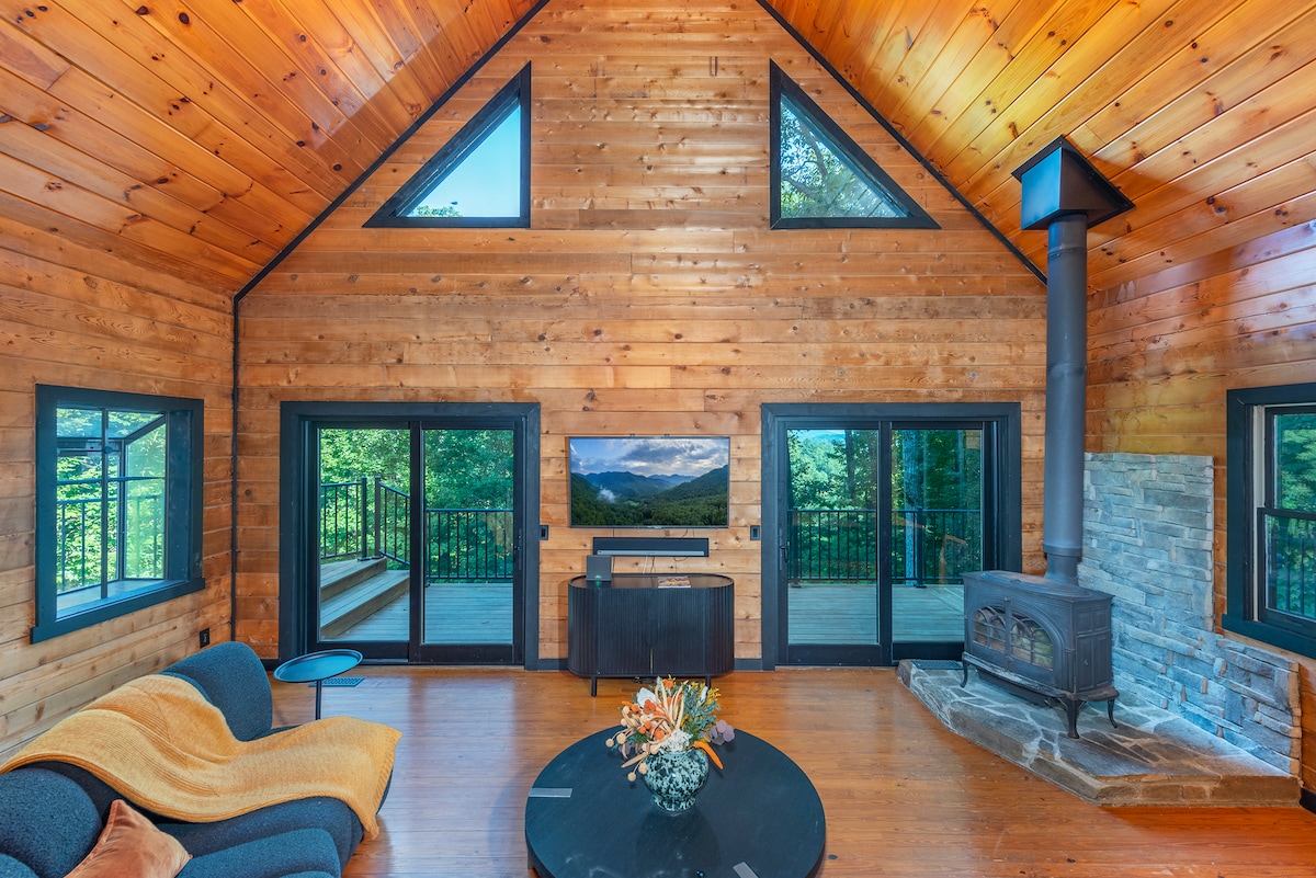 The living area features wooden walls and vaulted ceilings, complemented by large triangular windows that allow natural light to flood the space. A comfortable seating arrangement surrounds a coffee table with floral decor. A flat-screen TV is mounted on the wall, and a cozy wood stove provides warmth.