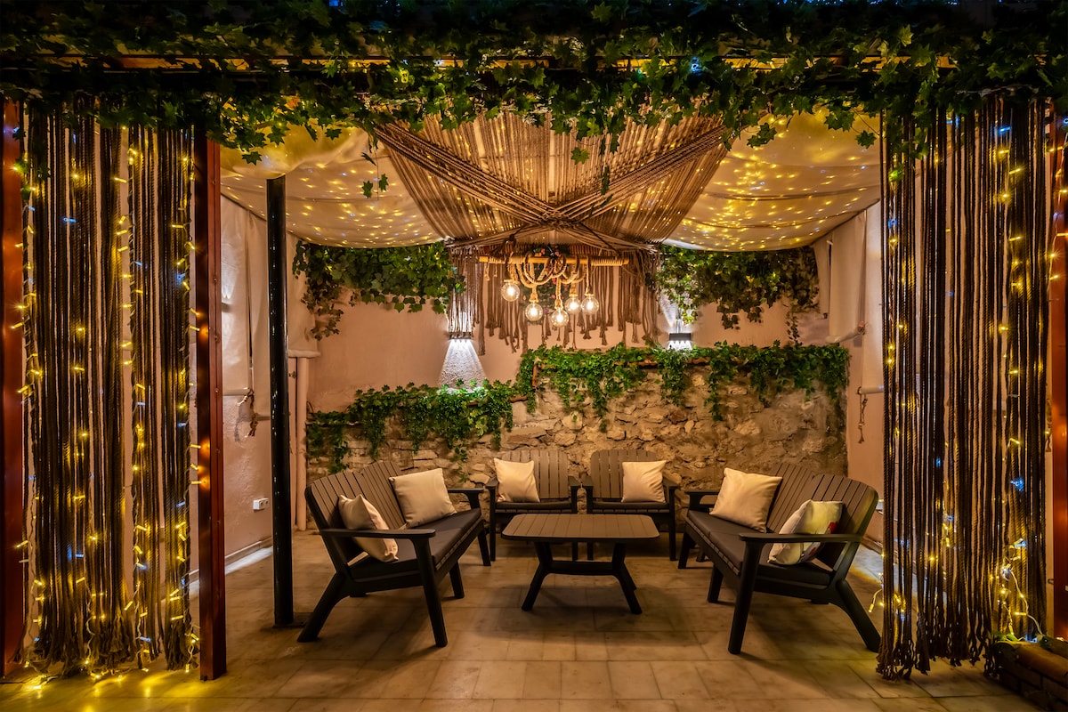 An outdoor seating area is surrounded by vertical greenery and soft glowing lights. Comfortable seating is arranged around a wooden coffee table under a decorative canopy, creating a unique social space. The atmosphere is complemented by warm lighting fixtures that add to the inviting setting.
