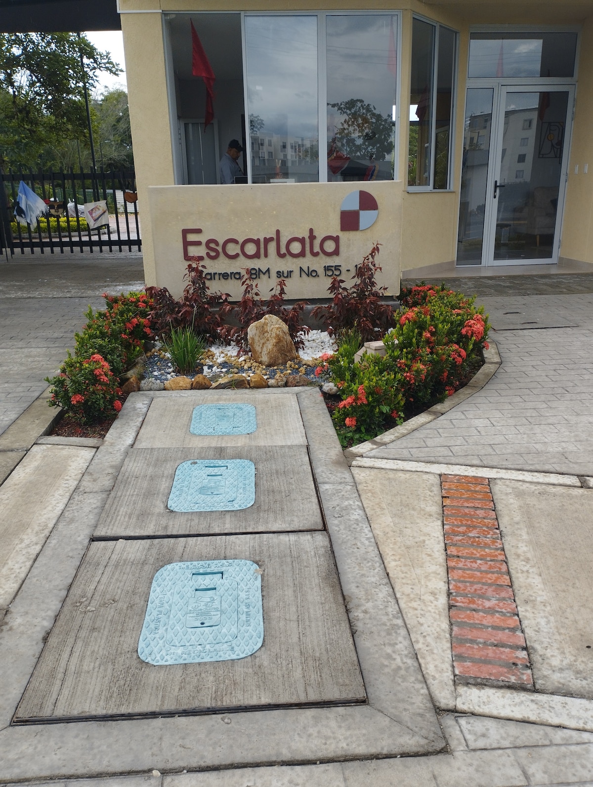 The entrance to the property showcases a neatly designed pathway lined with colorful plants and shrubs. A textured concrete surface leads towards a welcoming building facade, displaying the name 'Escarlata' prominently. The area is well-maintained, enhancing the overall approach to the apartment.