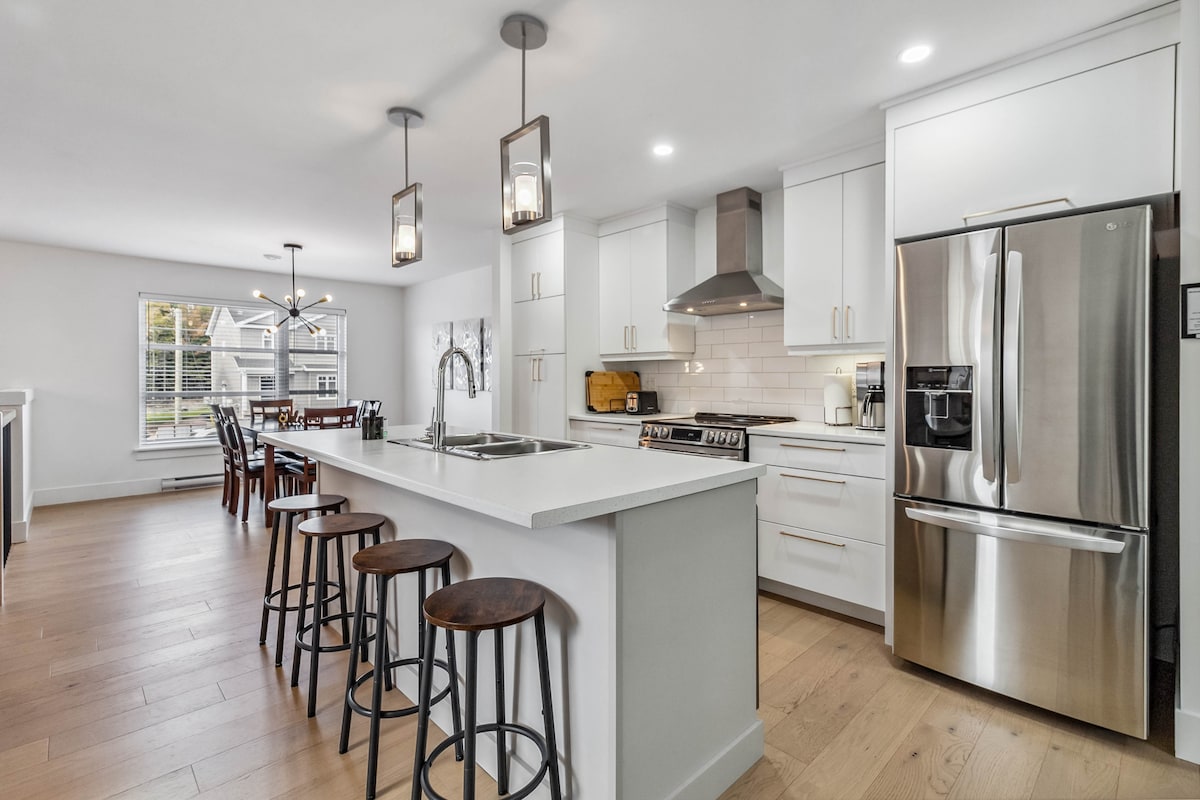 A modern kitchen is showcased with a central island featuring bar seating for four and stainless steel appliances. The open concept includes a dining area visible through a large window, illuminated by natural light. Sleek cabinetry and a stylish backsplash complete the inviting space.