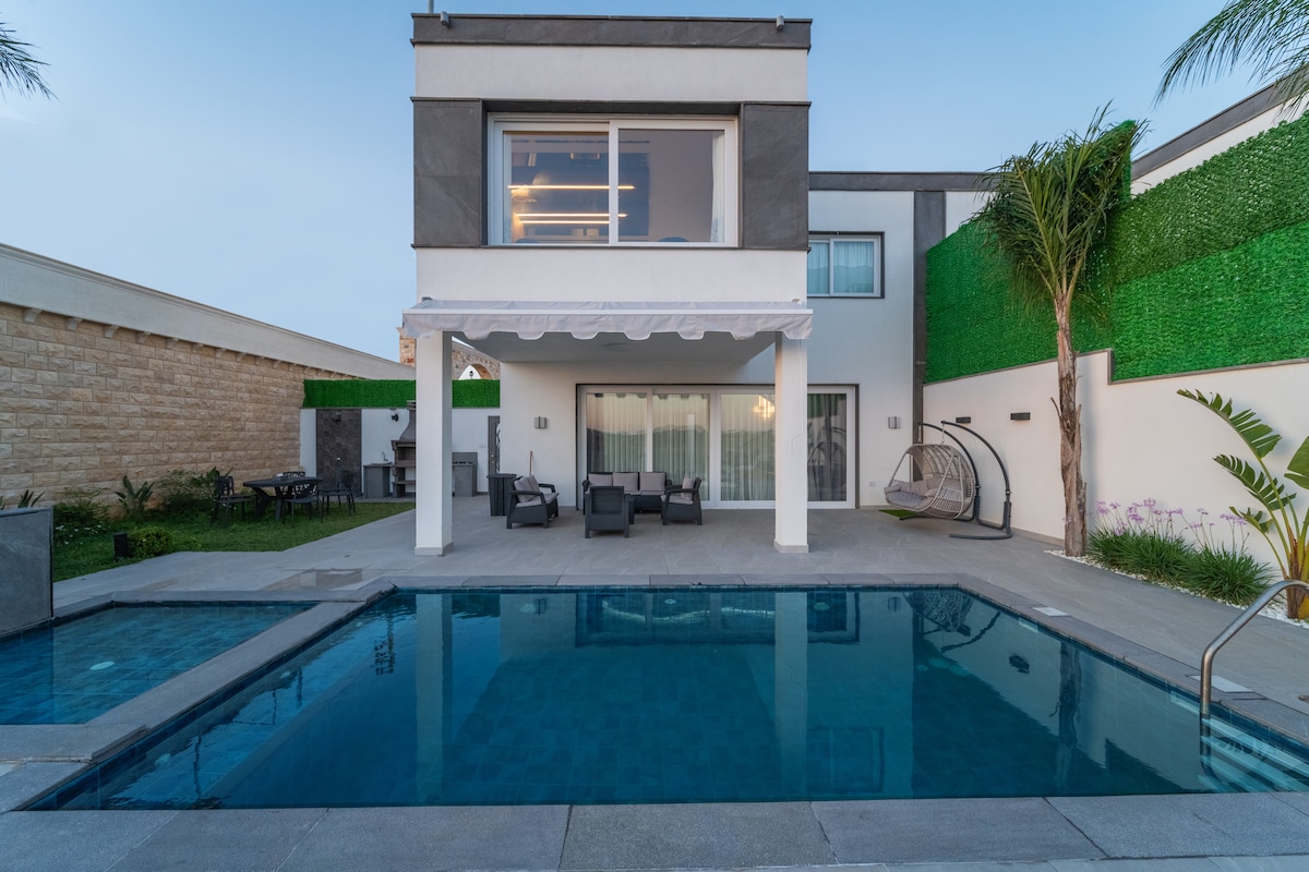 A modern villa features a private pool surrounded by a spacious patio. The expansive windows of the upper level provide a view of the outdoor seating area beneath a covered awning. Lush greenery lines the property, adding to the serene environment.
