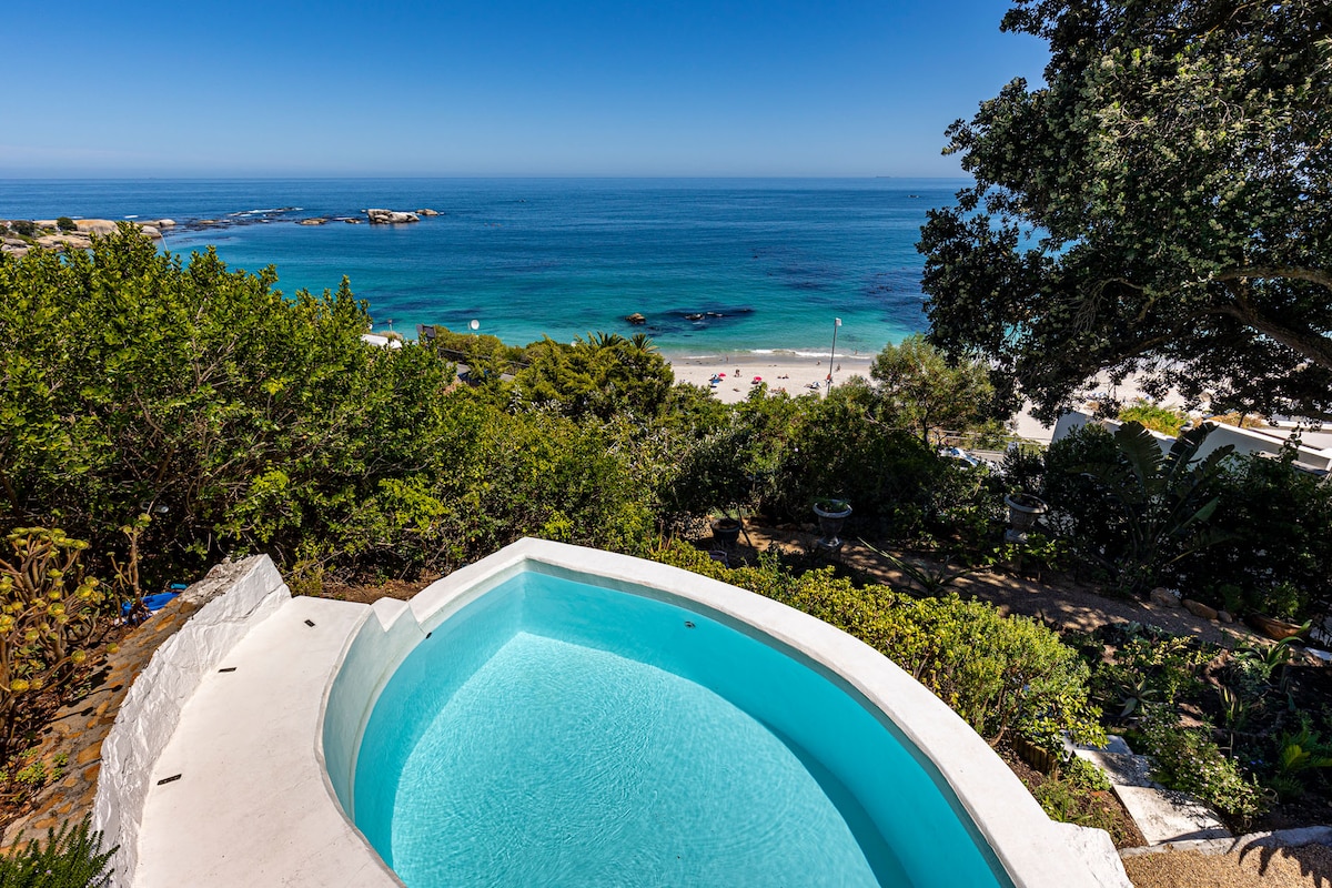 A curved swimming pool is framed by lush greenery, offering unobstructed views of the ocean and nearby beach. The serene blue water reflects the sky, creating a soothing atmosphere. The surrounding landscape adds to the natural beauty of this private outdoor space.