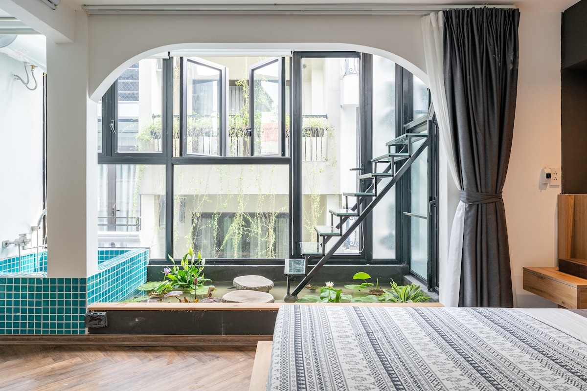 Large windows offer abundant natural light, overlooking a lush green area. A cozy sleeping space is visible with a patterned blanket atop the spacious bed. The contemporary design includes a small staircase leading to an upper level and a mini pool nearby.