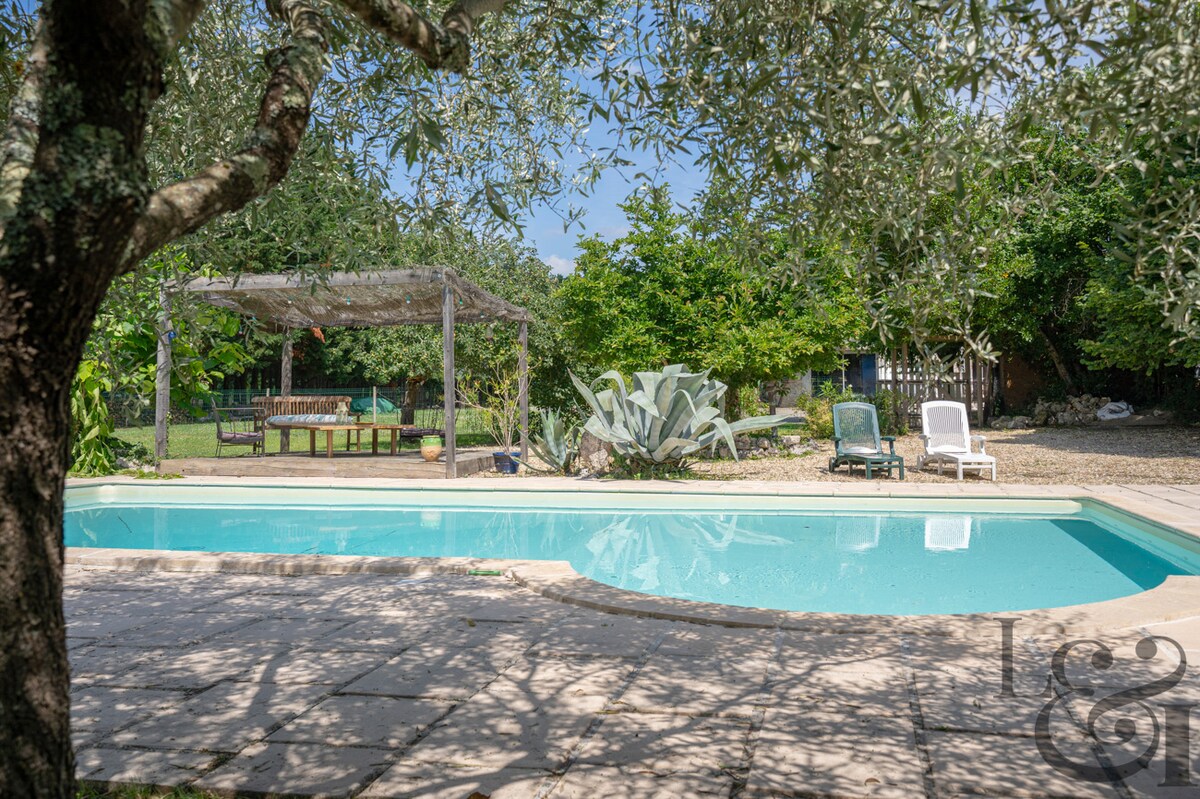 A tranquil swimming pool is surrounded by a stone patio, with lounge chairs positioned nearby. A shaded seating area is visible under a pergola, offering a peaceful space among the greenery of fruit trees and plants, enhancing the serene outdoor environment.