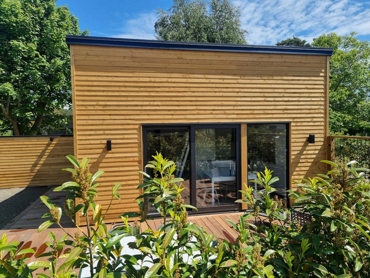 The exterior of the mini house features a sleek wooden facade with large glass doors framed by dark borders. The surrounding greenery adds a natural contrast, and the deck is partially visible, enhancing the inviting outdoor space.