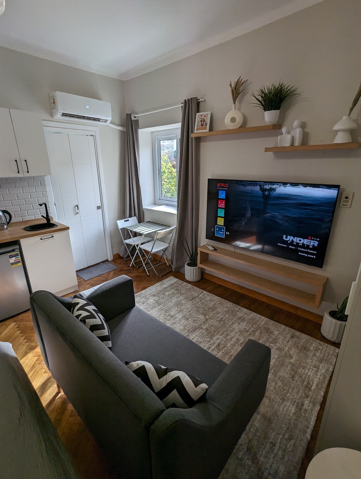 The studio features a compact living area with a gray sofa and a decorative rug. A mounted television is positioned above a wooden shelf, while natural light enters through a window. A small dining table is set against one wall, accompanied by two chairs.