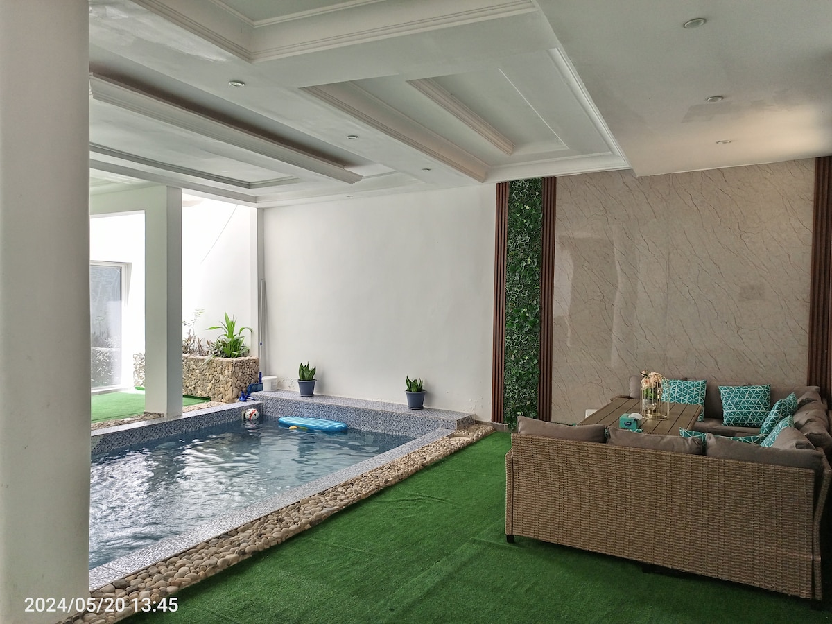 A serene indoor space features a covered private pool surrounded by a smooth stone border. Lush green plants are positioned along the walls, while a cozy seating area with turquoise-accented cushions provides a comfortable spot to relax. Natural light filters through the space.