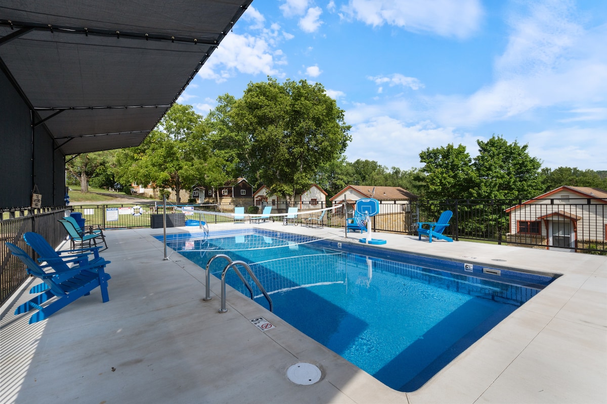 A refreshing outdoor pool area is displayed, featuring clear blue water surrounded by concrete decking. Several blue lounge chairs are positioned around the pool, while lush greenery and a few buildings are visible in the background, under a partly cloudy sky.
