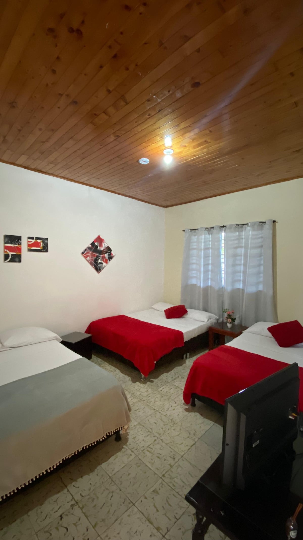 A bright room accommodates multiple beds covered with red and white linens. A mix of decorative wall art enhances the space. Natural light filters through sheer curtains, while a television is positioned on a low stand in the corner.